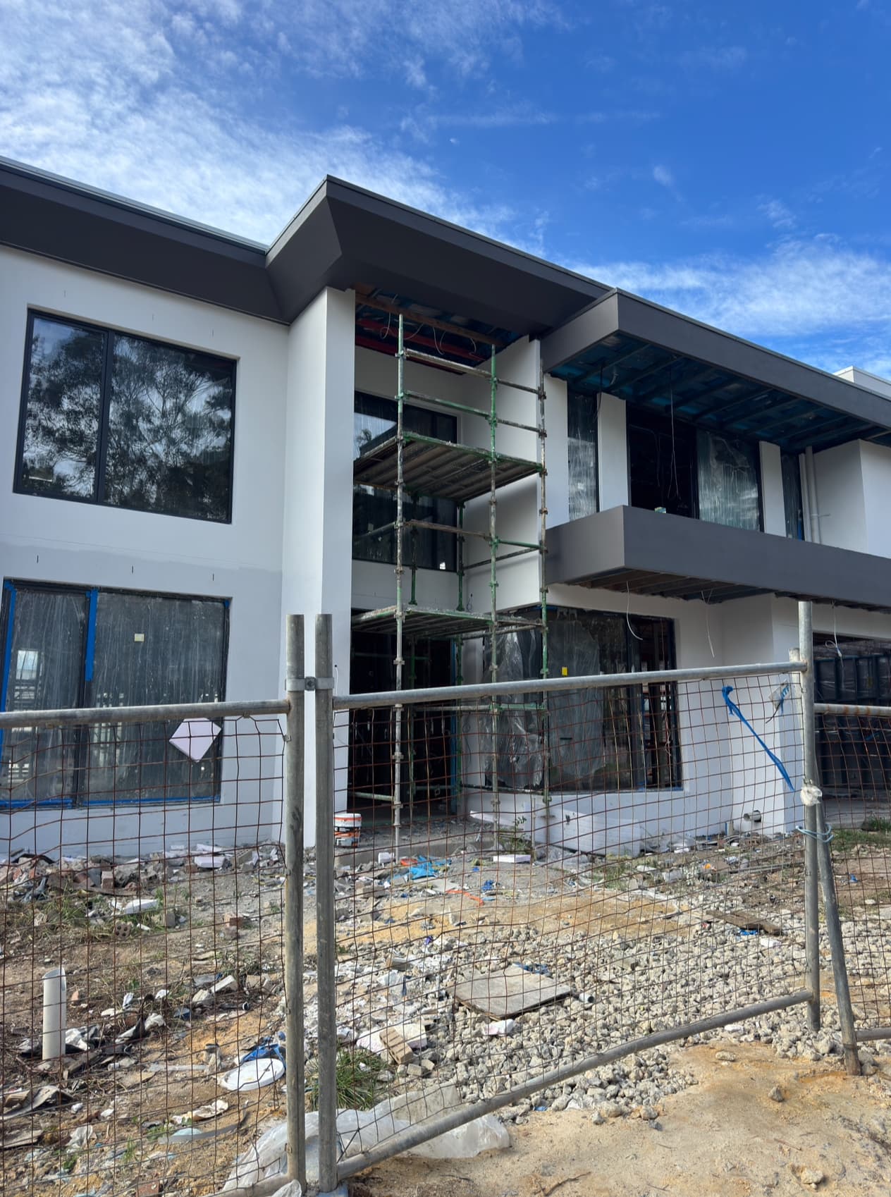 Modern white two-story house under construction with scaffolding and a wire fence in front.