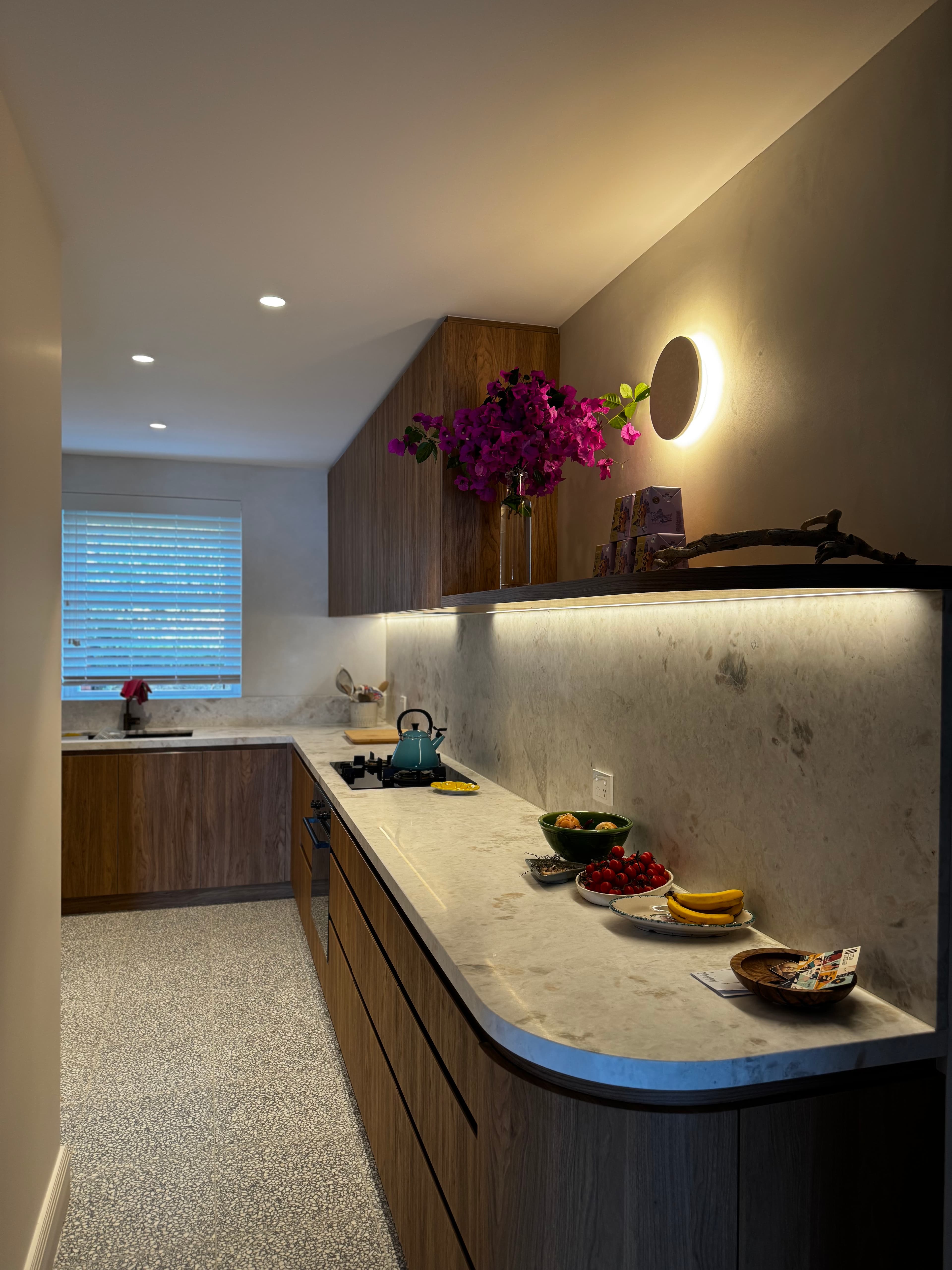 Modern kitchen with wood cabinets, stone countertops, terrazzo floors, and vibrant pink flowers.