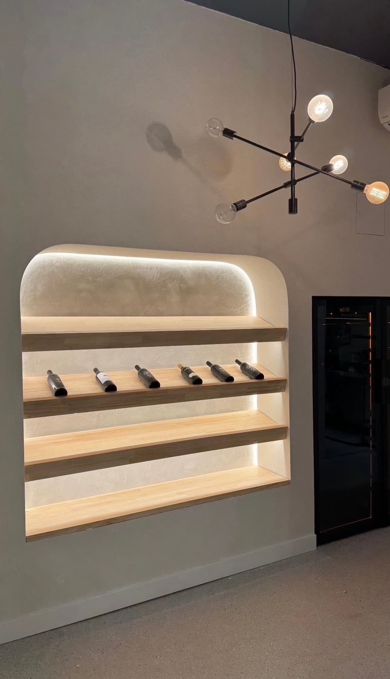 Modern backlit arched wooden shelves displaying wine bottles beneath a black sputnik chandelier.