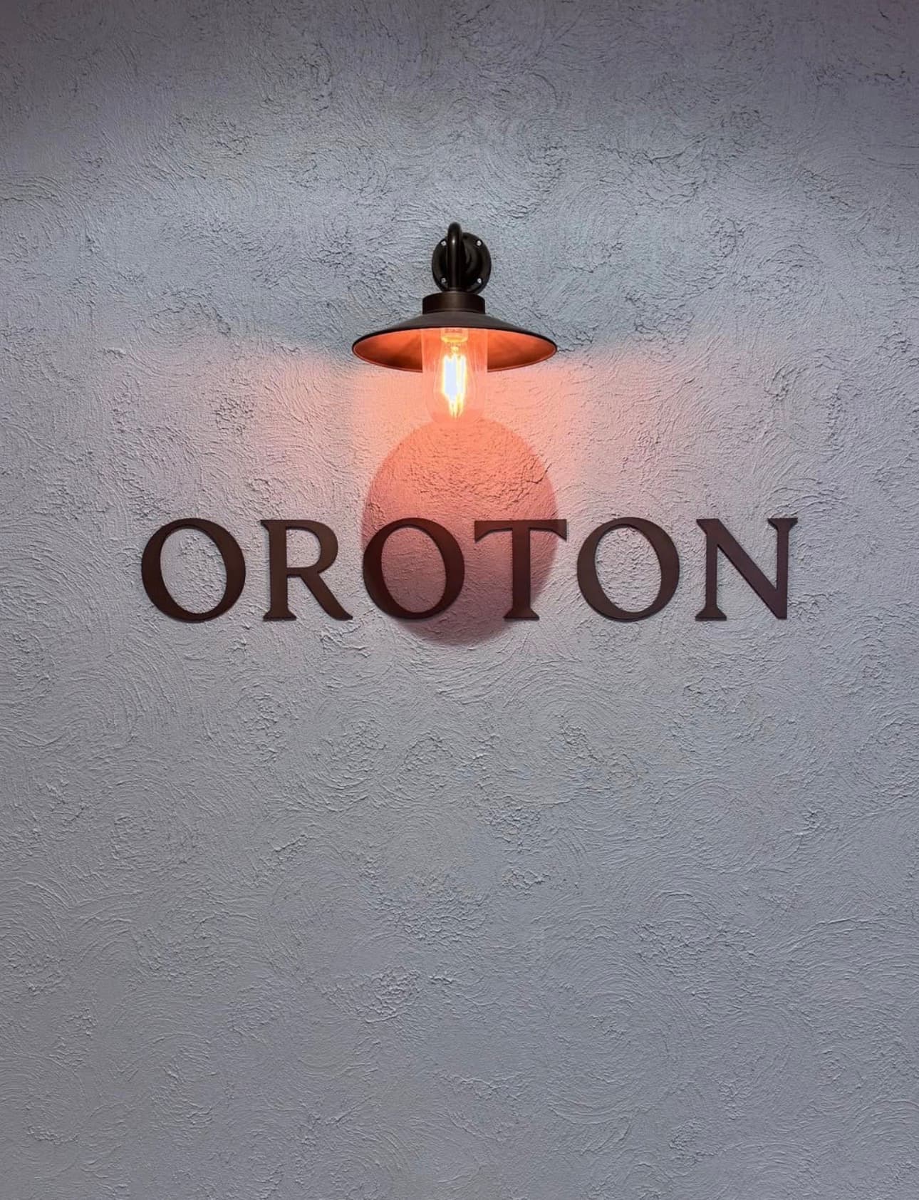 An industrial lamp illuminates the dark OROTON sign mounted on a textured gray wall.