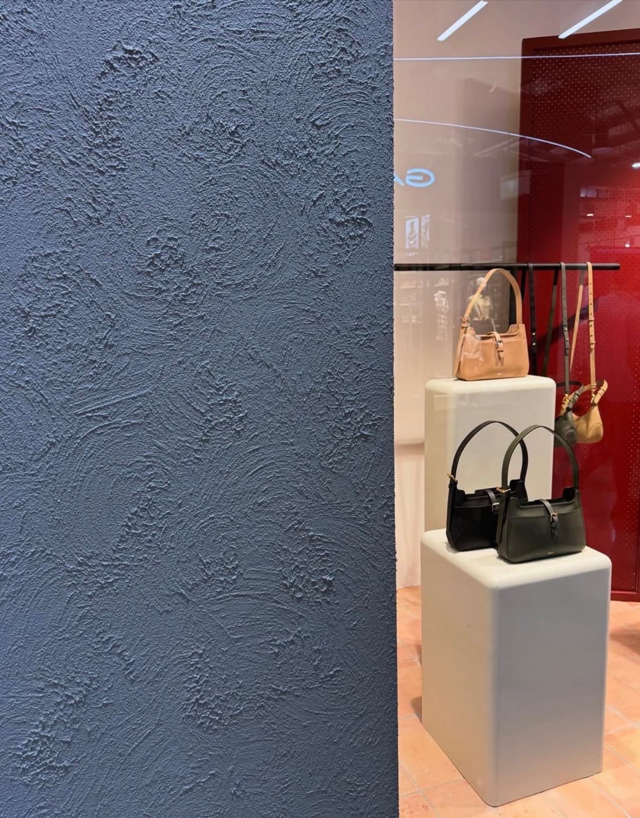 Textured blue wall framing a retail display of stylish handbags on white block pedestals.